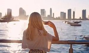 Nainen kaiteen edessä kuvamassa meristä kaupunkimaisemaa puhelimellaan