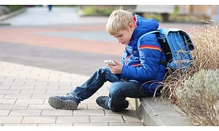 poika istumassa jalkakäytävällä reppunsa kanssa puhelin kädessään