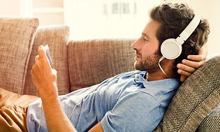 Mies rentoutumassa sohvalla puhelin kädessään kuulokkeet korvillaan