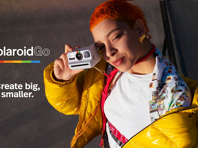Oranssitukkainen nainen keltaisessa takissa pitele Polaroid Go-kameraa ja teksti Create big, smaller.