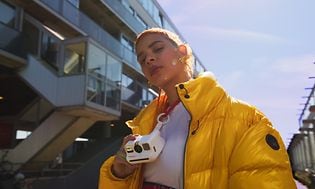 Nainen keltaisessa takissa Polaroid Go-kamera kaulassaan